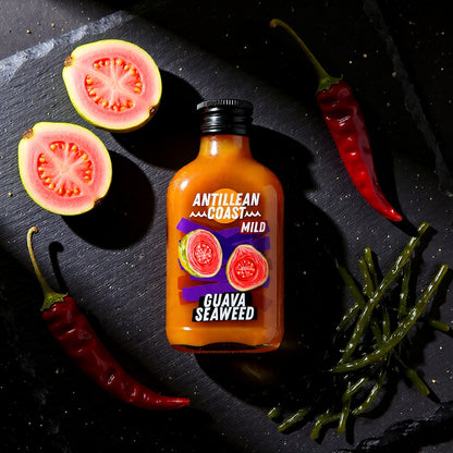 Guava Seaweed Hot Sauce Flasche auf Schiefer neben halbierten Guaven, roten Chilis und Meeresalgen
